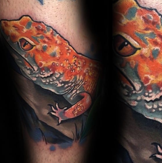 Vivid orange gecko tattoo with lifelike texture and shading, perched on a rock with dark tones and soft background details.