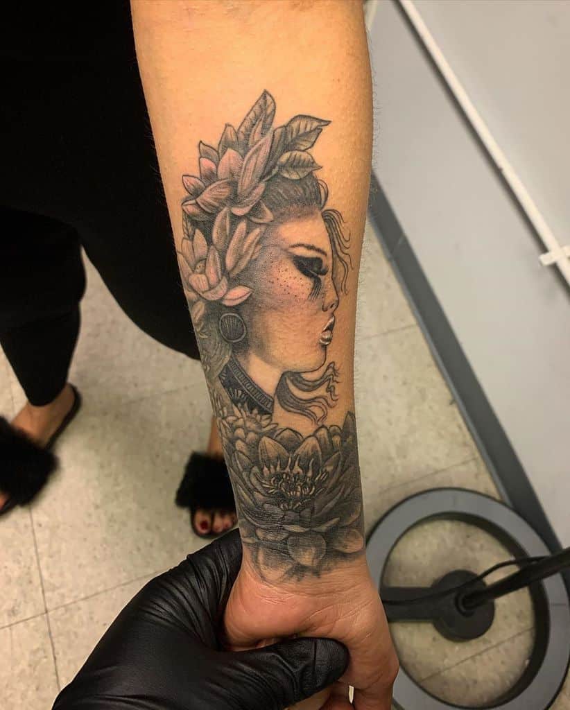 Black and gray forearm tattoo of a woman with a floral headpiece, delicate facial features, and a lotus flower at the wrist.
