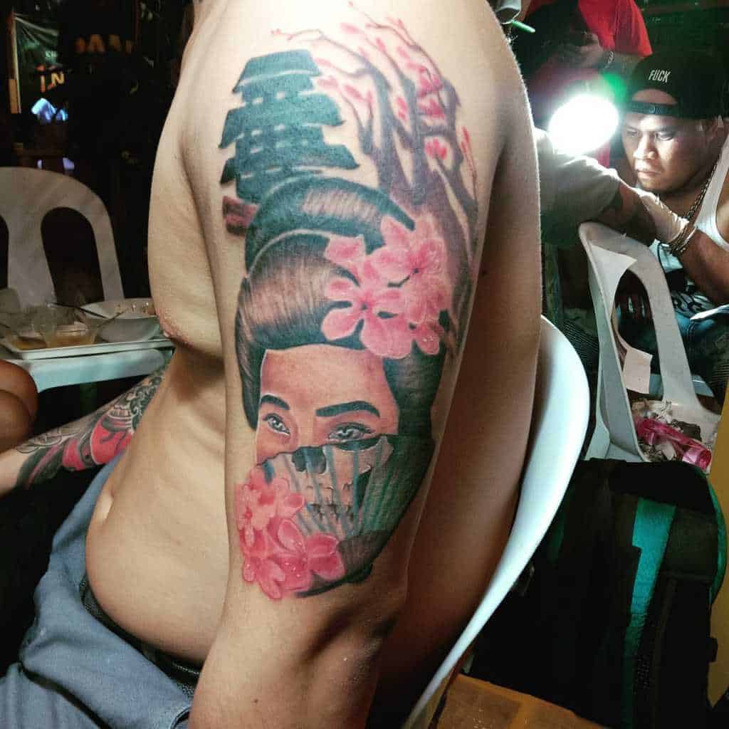 Bold upper arm tattoo of a Geisha with cherry blossoms, partially veiled face, intricate hair details, and Japanese kanji above.