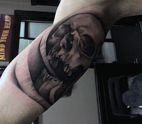 Black and grey tattoo of a melting skull within a circular frame, featuring smooth shading and a surreal, dark aesthetic on the bicep