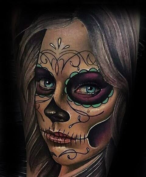 La Catrina tattoo of a woman with vivid blue eyes, colorful sugar skull makeup, and flowing hair in a realistic style.
