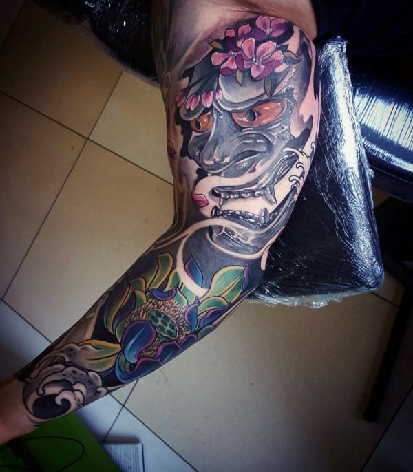 Japanese-style full sleeve tattoo featuring a fierce Hannya mask with glowing eyes, pink cherry blossoms, and a vibrant lotus flower