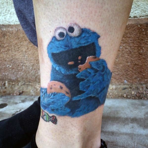 Fuzzy Cookie Monster tattoo on leg, joyfully holding and eating cookies with a wide smile and playful expression.