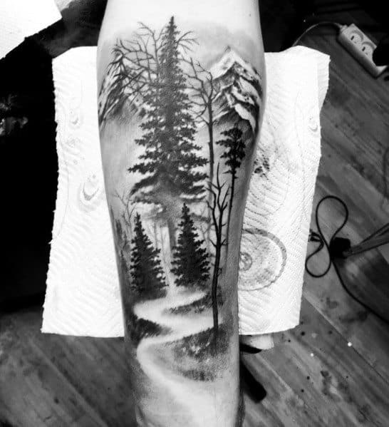 Black and grey forearm tattoo of a winding path through a dense pine forest, with misty mountains and bare tree branches in the background.