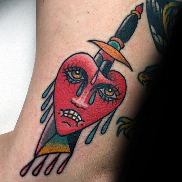 Tattoo of a stylized heart with a face, pierced by a dagger, with teardrops and blood drops
