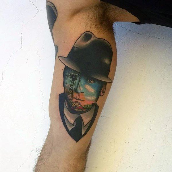 Surreal tattoo of a man in a suit and hat with a landscape scene replacing part of his face, blending portrait and dreamlike elements.