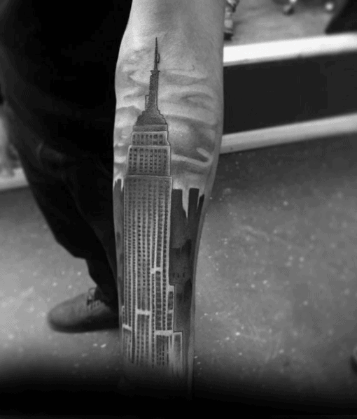 Detailed Empire State Building tattoo extending down the forearm, highlighted by realistic shading and a cloudy skyline backdrop.