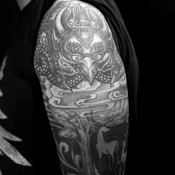 Garuda tattoo design with a detailed beaked face, cloud patterns, and a forest scene featuring a deer on the upper arm.