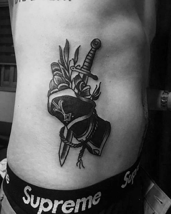 Black ink tattoo of an armored gauntlet holding a dagger and sword, with feather and rope details, placed on the side of the torso.