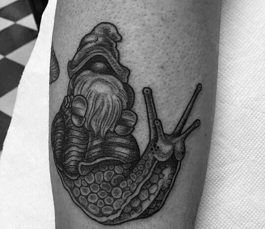 Black and white tattoo of a gnome with a long beard and a pointed hat, riding on the back of a detailed, textured snail.