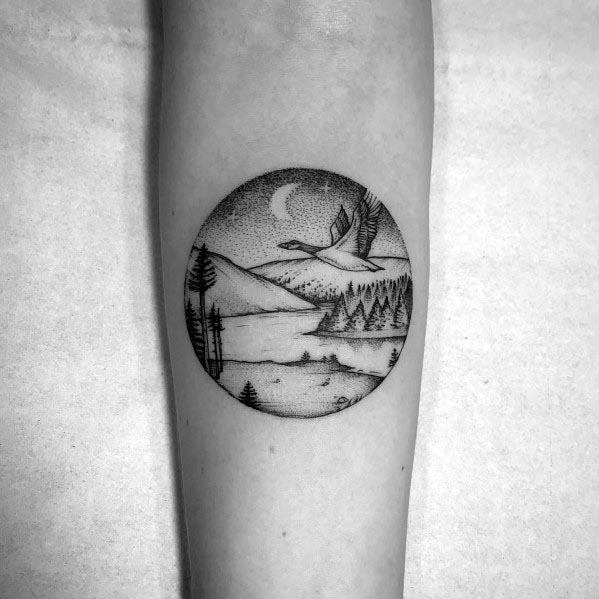 Black and white dotwork tattoo of a flying goose over a serene lake and mountain landscape, framed within a circular design on the forearm.