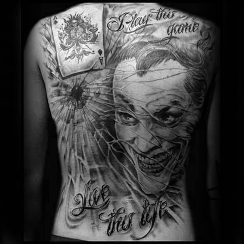 Full back tattoo featuring a sinister grinning face, shattered glass, playing card, and bold script reading “Play the game” and “Live the life.”