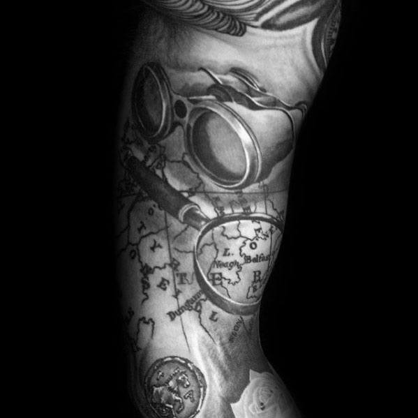 Tattoo featuring a magnifying glass over a vintage map, binoculars, and a coin, creating a realistic travel-inspired design with deep shading.