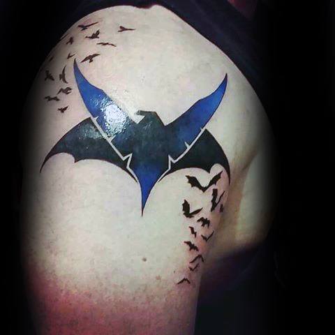Nightwing logo tattoo on the shoulder with a bold blue and black emblem surrounded by flying bats for a dynamic effect.