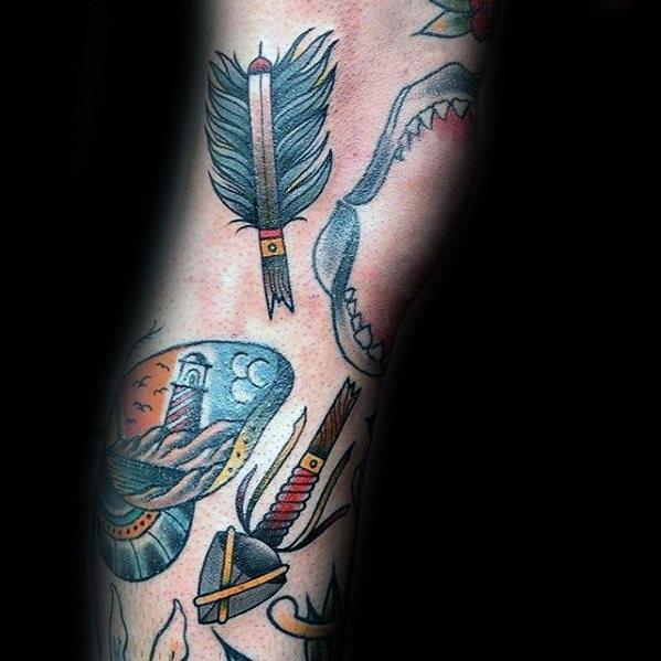 Broken arrow tattoo with a bold blue feather tip and red accent, placed among nautical and traditional motifs like shark jaws and a lighthouse.