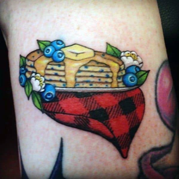 Traditional-style pancake tattoo with syrup, butter, blueberries, and flowers, resting on a red plaid heart for a cozy, rustic feel.