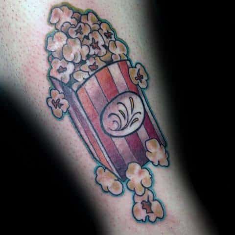 Tattoo of a tilted red-striped popcorn box spilling fluffy kernels, with a decorative swirl on the front of the container.