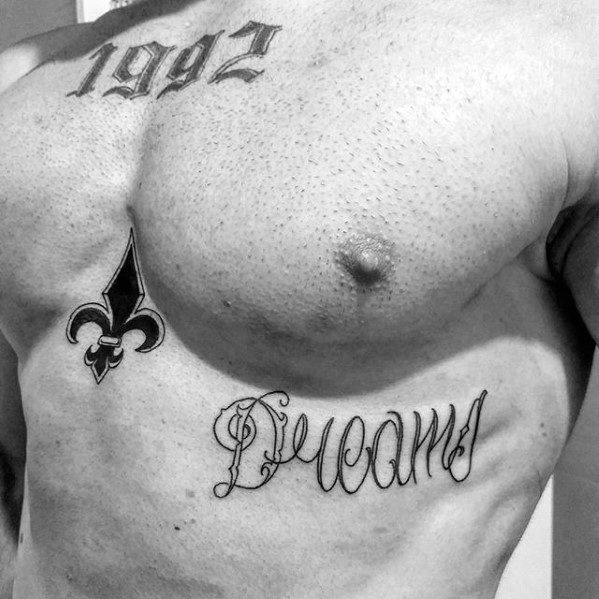 Chest tattoo featuring the word “Dreams” in ornate script, paired with a fleur-de-lis symbol and the year “1992” above.