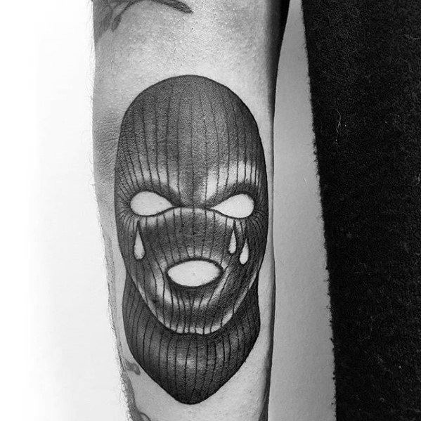 Tattoo of a black ski mask with hollow eyes and mouth, featuring three teardrops under one eye on the forearm.