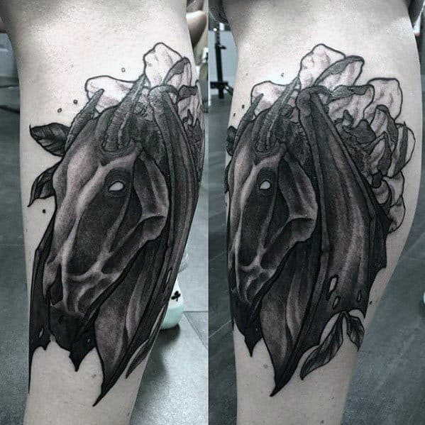 Dark Thestral head tattoo with flowing cloak-like wings and soft floral elements blending into the background on the leg.