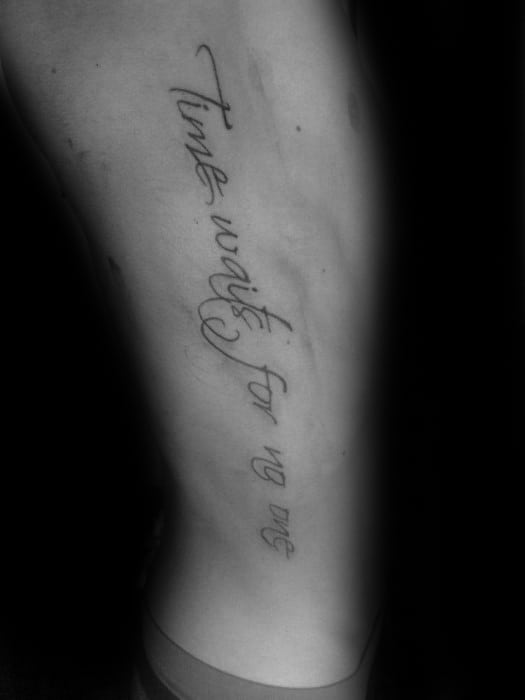 Minimalist tattoo in delicate script font reading Time waits for no one, placed vertically along the side of the leg.