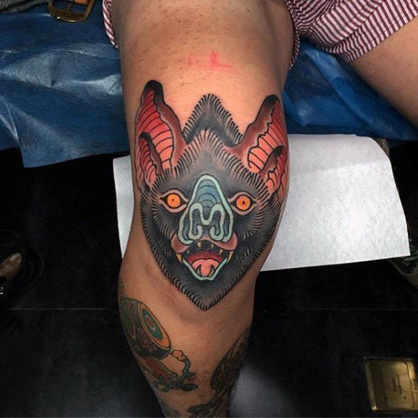 Bold neo-traditional bat head tattoo on the knee, with vivid red, blue, and orange details and sharp expression.
