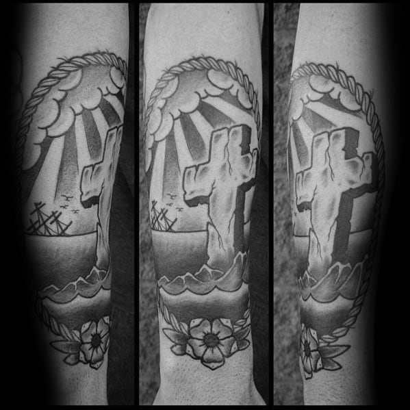 Black and grey tattoo of a stone cross on waves, with sun rays, ships in the background, framed by a rope border and floral design.