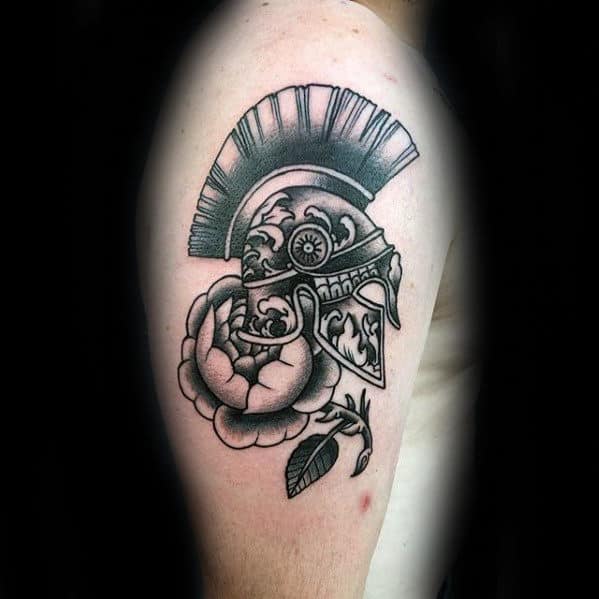 Arm tattoo of an ornate Spartan helmet above a blooming rose and leaves, in black and grey ink
