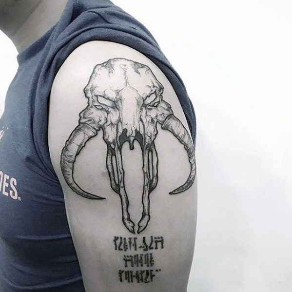 Detailed Mythosaur skull tattoo with long horns and Mandalorian script underneath, placed on the upper arm.