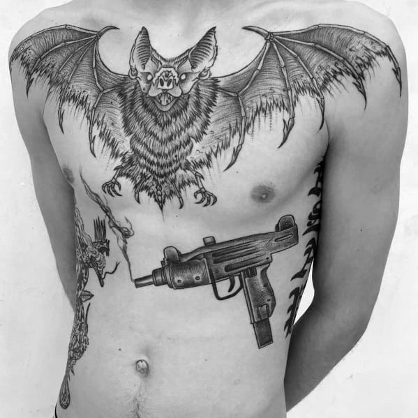 Black ink tattoo of an Uzi on the lower ribs, paired with a large bat chest piece. Dark, detailed, and gothic-inspired design.