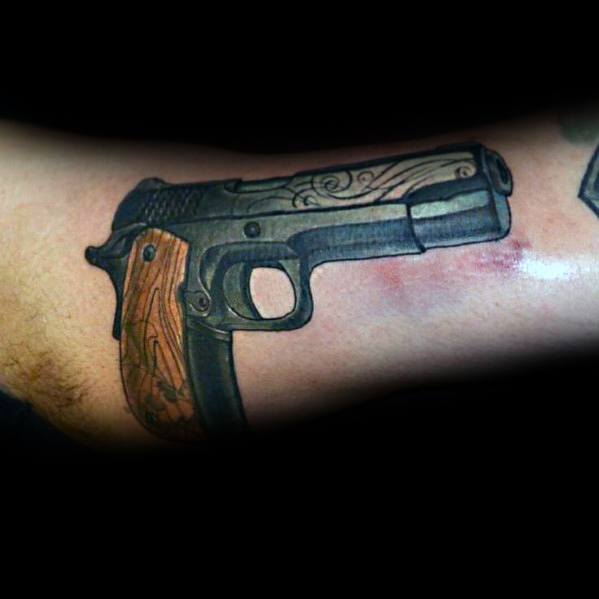 Realistic tattoo of a 1911 pistol on the arm, featuring engraved details and wooden grips, designed with bold shading for a three-dimensional effect.