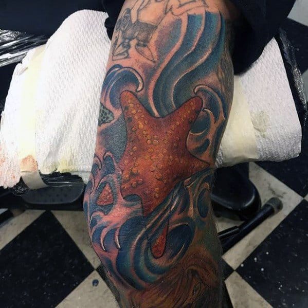 Large orange starfish tattoo on the leg, surrounded by bold blue ocean waves and detailed shading in a dynamic underwater scene.