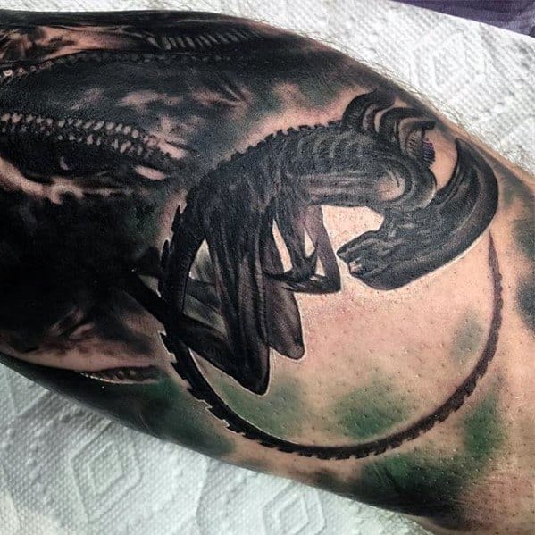 Silhouetted Xenomorph tattoo in a crouching pose with a long curved tail, dark shading, and green background highlights.