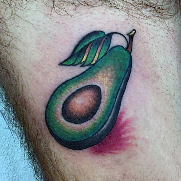 Colorful avocado tattoo with a stylized leaf on top and bold shading, placed on the thigh with a soft pink background blush.