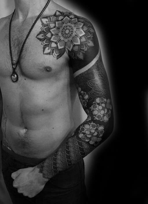 A black and grey full sleeve tattoo featuring a large mandala design on the shoulder, bold geometric patterns along the arm, and detailed shading with a solid black armband near the bicep