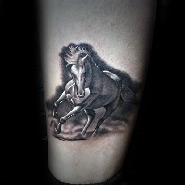 Realistic black and grey tattoo of a galloping horse with flowing mane and dust clouds, placed on the lower leg