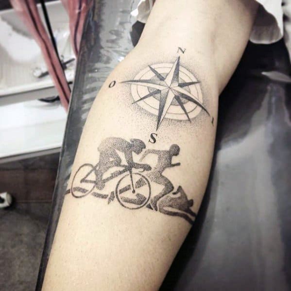 Tattoo featuring a compass design with north and south markers, intertwined with a depiction of an Ironman race sequence: swimming, cycling, and running.