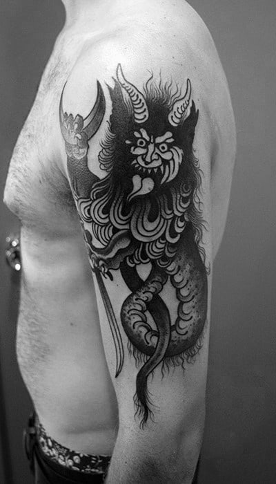 Bold black Krampus tattoo on upper arm showing a fierce, horned creature with clawed hands, swirling fur, and a long tail.