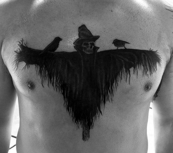 Chest tattoo of a shadowy scarecrow with a skeletal face, wide straw arms, two perched birds, and a tattered silhouette.