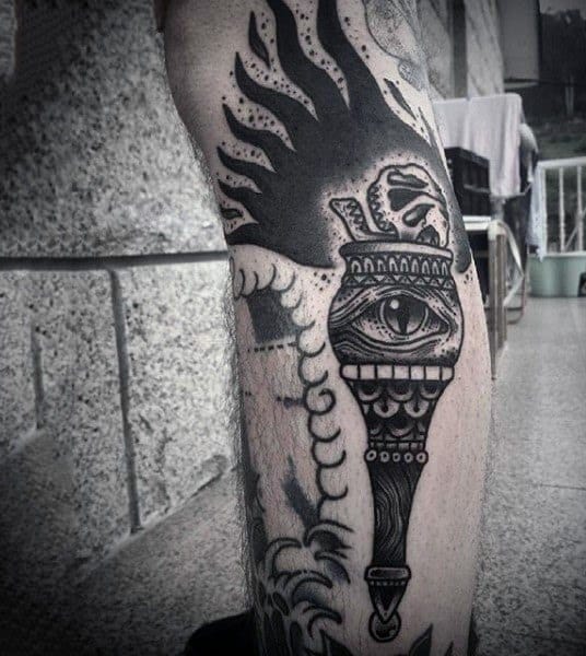 Dark torch tattoo with intense blackwork, featuring an all-seeing eye in the center and bold flame shapes rising above.