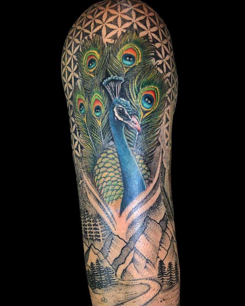 Colorful peacock tattoo on the arm with vibrant blue feathers, surrounded by peacock tail feathers, geometric patterns, and a landscape with mountains and trees.