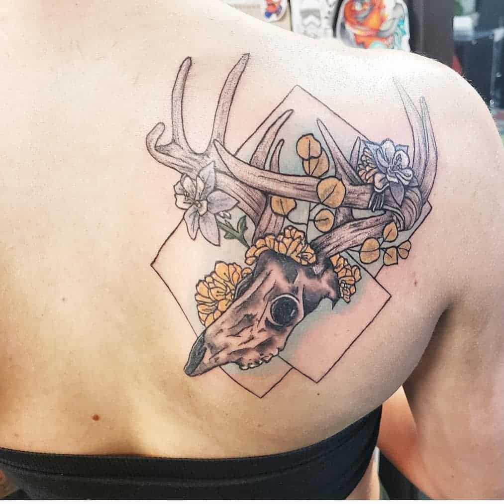 Tattoo on upper back of a deer skull with antlers, surrounded by flowers in a geometric shape