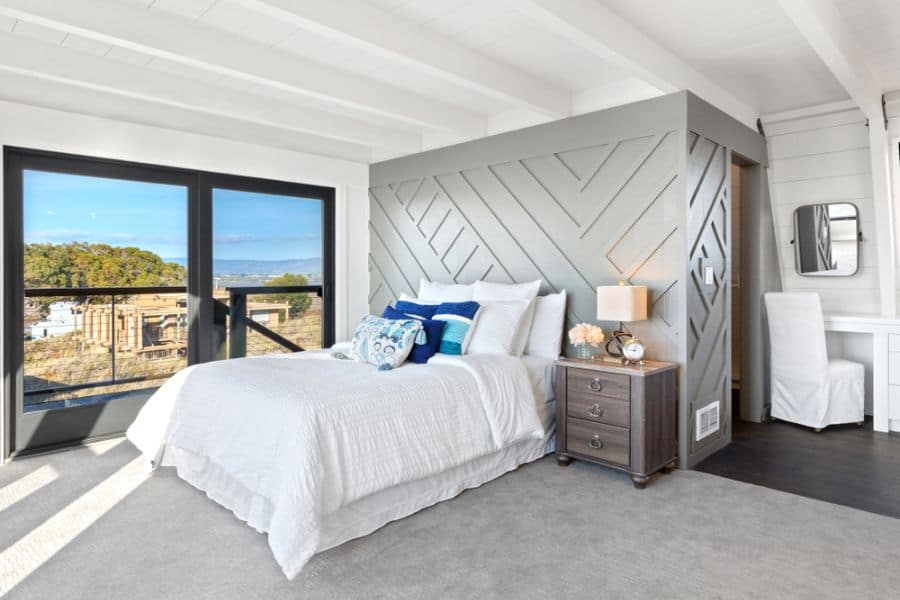 Bright bedroom with a geometric accent wall, large windows, white bedding, and a wooden nightstand with a lamp