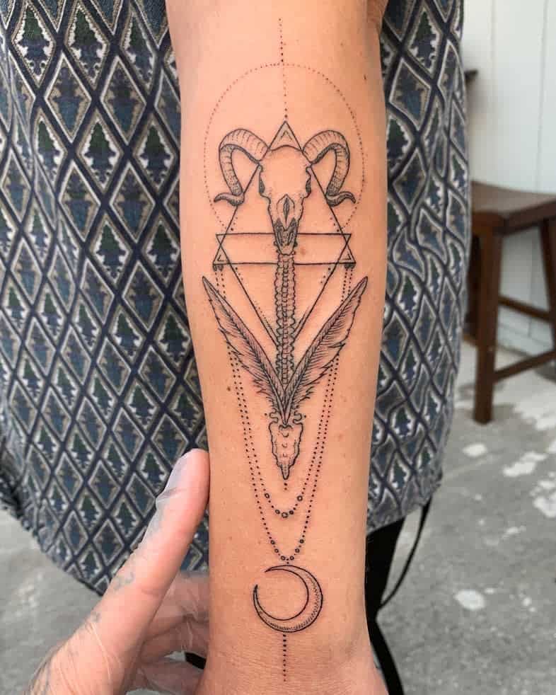 Forearm tattoo of a ram skull, geometric shapes, feathers, and a crescent moon design