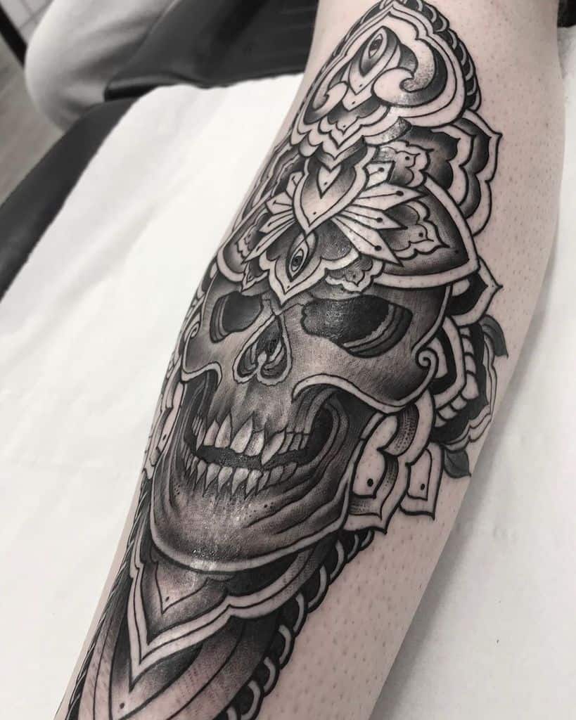 Tattoo of an intricately detailed skull with a third eye, surrounded by ornate mandala patterns, inked in black and grey shading on the forearm.