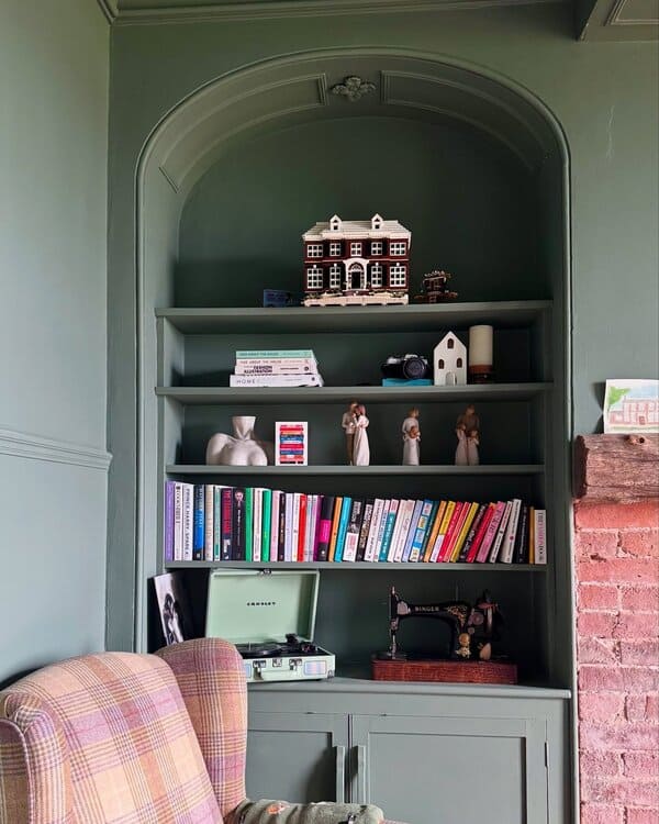 Green alcove shelving with books, a miniature house, vintage sewing machine, and a plaid armchair beside an exposed brick fireplace.