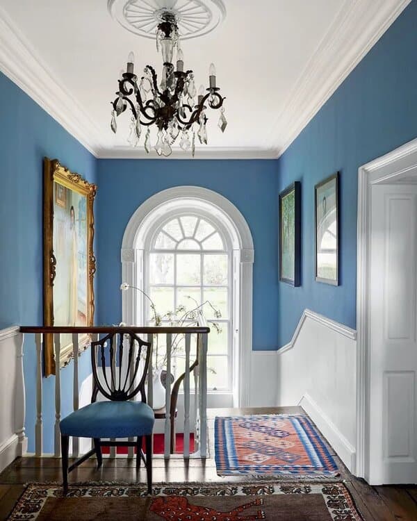 Bright blue hallway with white wainscoting, an arched window, a vintage chandelier, and patterned rugs for a stylish, classic look.