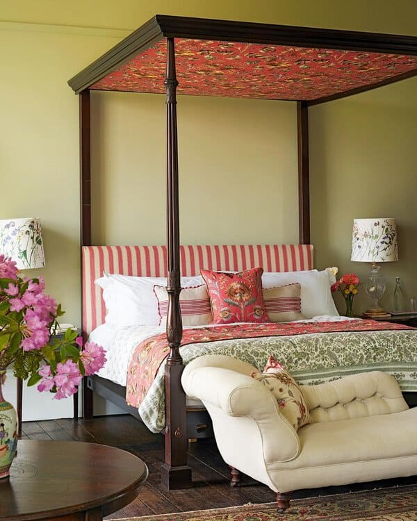 A four-poster bed with a red floral canopy, striped headboard, colorful pillows, and a white armchair with a floral vase nearby.