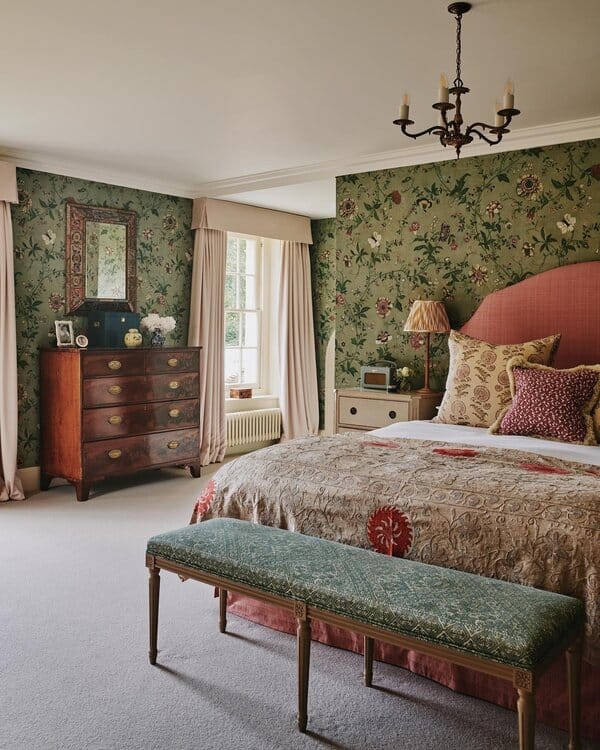 Bedroom with green floral wallpaper, pink upholstered headboard, antique chest of drawers, and a vintage chandelier for a garden-inspired feel.