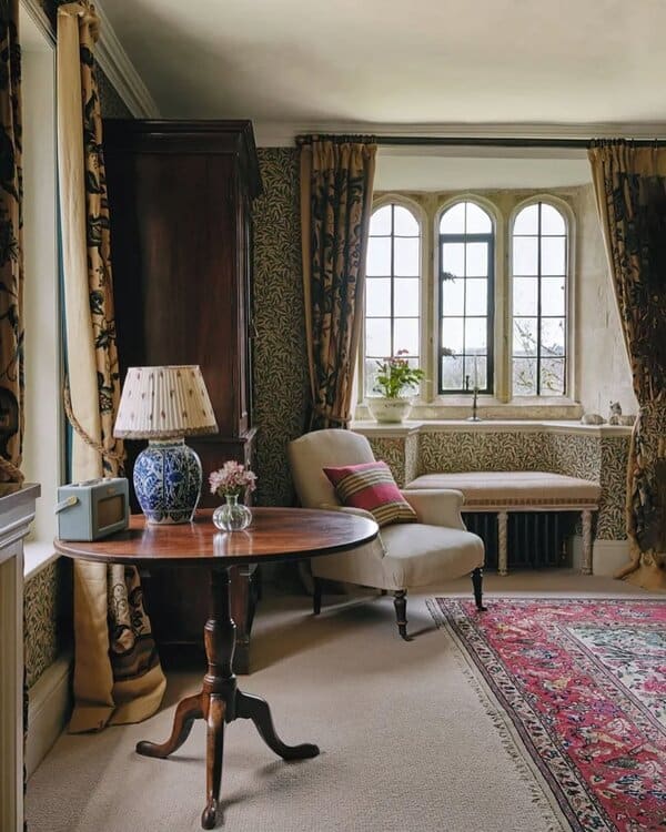A cozy corner of a Georgian-style room featuring a window nook, antique furniture, and a decorative table with a lamp and flowers.